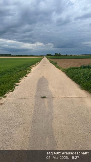 Auf einem Feldweg mit Betonplatten, zwischen denen Gras wächst, sieht man den Schatten eines Radfahrenden. 

Der Himmel ist bewölkt.