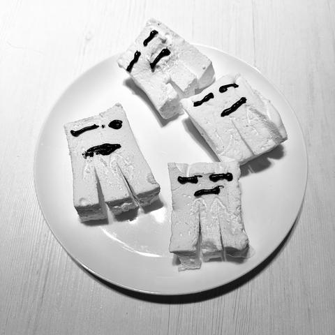 Ein weißer Teller hält vier Dessertstücke, die verspielten Geistergesichtern ähneln und aus einer weichen, marshmallowähnlichen Textur geformt sind. Jedes Stück zeigt karikaturhafte Ausdrücke aus dunklem Schokoladen-Nieselregen. Das Bild ist in Schwarzweiß.
