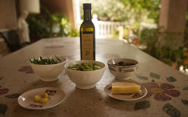 On a stone table in the garden a bottle of olive oil and several bowls and plates with ingredients are arranged. These are clockwise from top left: rocket, broad beans, toasted almonds, parmesan and garlic.