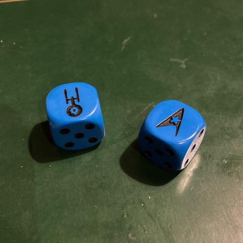 Two blue dice, on the face of one is the silhouette of a Constitution class Starship, and on another the symbol of the Enterprise crew, later adopted by Starfleet and used in their communicator badges.