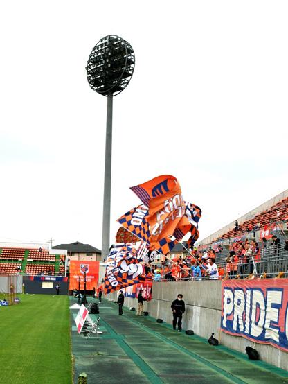 In the home end supporters in the standing area wave huge flags in the club's colors orange and blue. I captured them from the side and the large stadium lights pole also made the shot. The sky is gray making the orange colors pop.