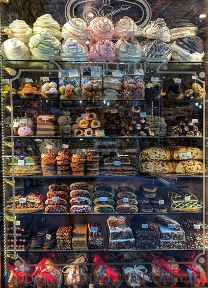 A brightly lit shop window display of "La Bottega Del Pasticcere" showcases multiple shelves filled with a variety of pastries, cakes, and chocolates. The top shelf features meringue-like sweets in various pastel colours. Below, several shelves display an assortment of small cakes, pastries with fillings, and what appear to be chocolate truffles or similar confections. The bottom shelf contains larger slices of cake and chocolate treats, some wrapped with red ribbons. The overall impression is of a tempting and well-stocked patisserie.