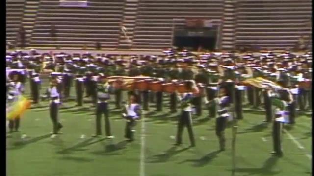 A drum corps, the Cavaliers, performing on a football field. Members are dressed in coordinated uniforms, holding instruments and flags, with stadium seating in the background.