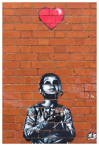 A red brick wall with a black and white murale, probably a canvas attached to the wall, representing a boy with a dreamy sight looking above himself to a bright red heart with a black contour. In the bottom right corner a white mark that could resemble the artist signature.