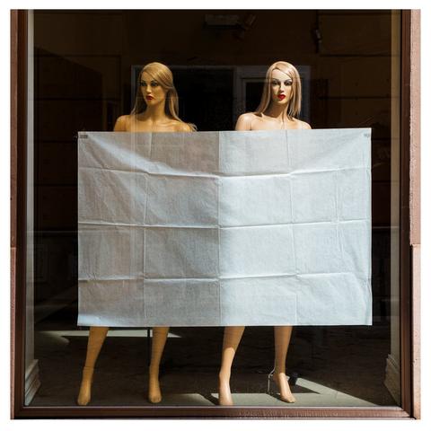 Zwei (nackte) Schaufensterpuppen stehen nebeneinander in einem Schaufenster. Die Puppen haben blonde Perücken und sind auf einer glatten Fläche platziert, die von Tageslicht durchflutet wird. Vor den beiden Puppen hat jemand ein Stoffstück gehängt, das den Blick versperrt.