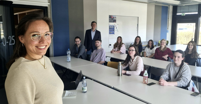 Credit: Hochschule Hof Alt-Text: Katharina Klug vor Studierenden in einem Seminarraum der Hochschule Hof, alle blicken lächelnd in die Kamera. / Katharina Klug with students in a seminar room at Hof University, all smiling at the camera.