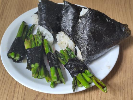 On a white plate, three onigiri triangles are resting on each other. In front of them are four bundles of fried asparagus, each bundle wrapped in a small strap of nori.