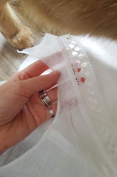 A close up photo of the absolutely tiny rolled hem going on the under arm scoop of a little top, in sheer and fine white linen. It's so thin Sini's fingers are fully visible through it. There is a medium to narrow strip of off white linen lace sewn onto the front part on a diagonal, the seam allowances of it finished with tidy machine stitching. The side seam has been felled by hand on both sides of the seam allowance, making it very flat. A sewing needle with linen thread is hanging onto the hem in progress, Sini's pink hand holding onto the sewing. The back leg and belly of a ginger tabby cat dominates the top of frame, as he is traipsing over the sewing at moment of photo.