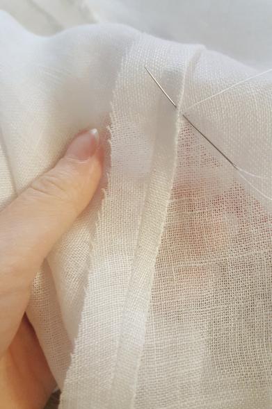 The sewing on the side seam, inside. A 1.2cm seam allowance has been folded and rolled over in such a way that all raw edges are tucked away, hidden and secured, and that is then sewn on with tiny whipstitch. The right side is being sewn, the left side still has the raw seam allowance out. Sini's fairly bloodless thumb holding onto the sewing, rest of the fingers partially visible through the sheer fabric.