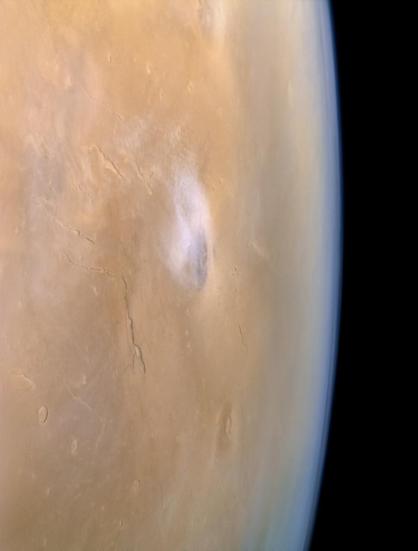 A view of Mars from orbit, captured by NASA's Viking 1 in 1978, showing the planet’s rusty reddish surface curving toward space at the right edge. In the center, a bright, whitish, oval shaped cloud covers the peak of Elysium Mons, a large volcano. To the left of it, dark, fractured cracks run across the surface. These are the Elysium Fossae near the top and Elysium Chasma below. At the bottom of the image, Albor Tholus appears as a subtle, circular volcanic rise. Near the top, Hecates Tholus is faintly visible as a smaller, rounded volcano. The background to the right is the blackness of space, creating a stark contrast with the soft Martian atmosphere fading into a bluish haze near the edge, which is one of the most stunning aspects of the image.