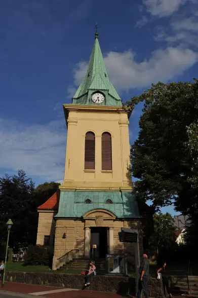 The image appears to depict a building with a church-like architecture, featuring a tall spire with a green roof, a clock near the top, and an entrance at the base. The surrounding area includes trees and a pathway, suggesting it's part of a public or outdoor space. The sky is partly cloudy, indicating a clear day.

Image Credits: Wikimedia / Hannes Grobe / CC BY 3.0