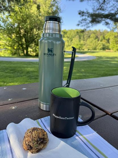 The after-walk payoff this morning, was a breakfast cookie from WildFlour Bakery in Saluda, NC (from the freezer) and some coffee, sitting on a picnic table under some very large pine trees, with a view of one of the paths, and the morning sun lighting up nearby and farther-away trees.

The cookie dough was clearly scooped out with an ice cream scoop and then baked, but it's so full of goodies that it doesn't flatten at all.  They cooked it long enough this time, too, sometimes they're a bit gummy.

The table is "set" with a white and blue striped towel, and holds a black coffee cup with a green interior, labeled with the term "#AppellateTwitter" from my days of stalking those folks on the bad place.

A green Stanley thermos is waiting behind the mug, to provide several refills.  It was about 55F when we got to this point of the morning, just about perfect.
