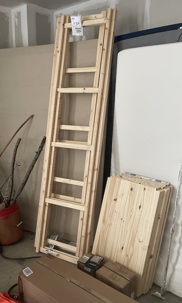 A picture of bare wood shelving. The side panels look like ladders and go up just past eight feet. The wood shelves themselves are stacked against the wall. The wood is untreated and unstained.