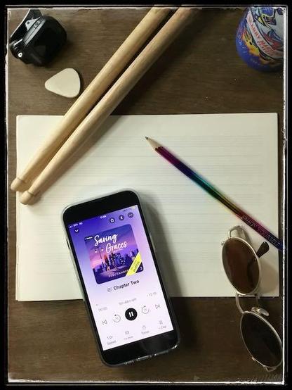 On a wooden background, an iPhone with the cover of Saving Graces by Ruby Landers, narrated by Quinn Riley, on music paper, next to drumsticks, a guitar pick, a rainbow pencil and sunglasses.