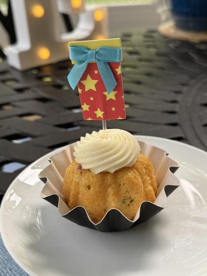 A confetti bundtini with a package flag sticking out of the top sits in its fluted cup on a small white plate on a black trellis table.  Much is out of focus behind the cake, but part of a lighted capital "E" is visible at the top left of the photo.

The little bundt cake is vanilla with various bits of colored whatever interspersed in the cake, then topped with a ridiculous amount of butter cream frosting.  And a red "package" made of paper with stars on it, and a tiny blue ribbon on a clear plastic toothpick, is topping the little morsel.

No, this is not the only bundtini.