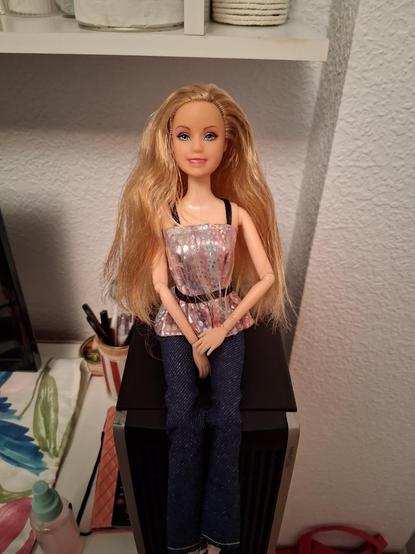 La muñeca sentada encima de mi PC con unos vaqueros, unos tacones plateados y un top brillante plateado y rosa con una cintura y tirantes negros