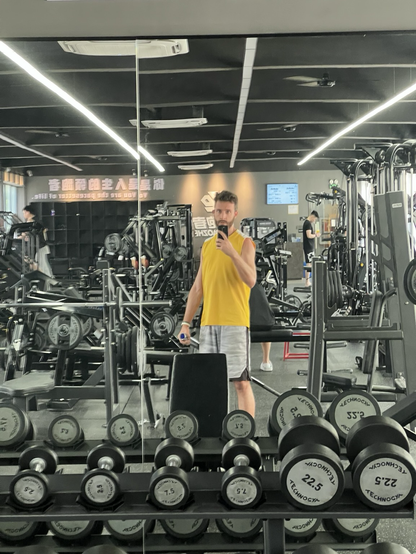 A man takes a selfie in a gym mirror. His other hand holds a barbell. In front of the mirror is a rack of dumbbells