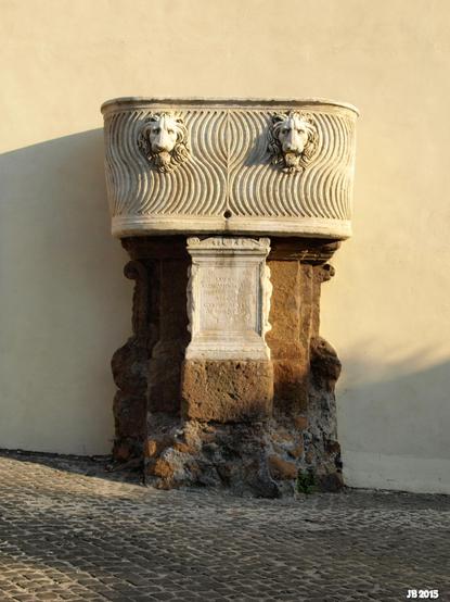 Farbbild eines alten Brunnens in der Kleinstadt Zagarolo in Lazio, Italien. Auf einem massiven gemauerten Unterbau steht eine Wanne aus hellem Stein mit symmetrischem bogenförmigem Muster und zwei sehr vermenschlichten Tierköpfen.