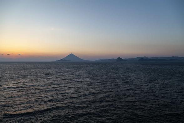 It's evening, just after sunset at sea with the horizon marked by a pale, narrow band of distant cloud glowing orange at its upper edge before fading through light gold and white to a dusky blue of clear sky as it reaches up to the top of view. The sea is calm, rippled, and inky black now that sun is below the curve of the Earth. Land stretches across the view of the horizon from the right about two thirds of the way. It's mostly low land with rises and falls of a hilly landscape in a couple of shades of grey-blue silhouette, but there's a volcanic mountain peak at the far left, almost perfectly triangular, its very top brushing the orange-gold band of glowing sky above the far off cloud line. It feels like a warm day that's suddenly cooled but it's quiet and relaxing.