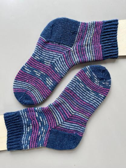Pair of had knit socks on blockers. The have shallow stripes in pinks and light blue paired with dark blue and solid cuff, heel and toe in dark blue. They are done with a technique called helical knitting.