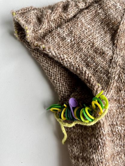 Armhole of an unfinished knitted cardigan. Most stitches are on waste yarn, but on the bottom part a series of stitch markers has been inserted to indicate where to pick up stitches.