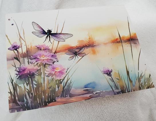 A greeting card depicting a serene lakeside scene. In the foreground are clusters of soft purple daisy-like flowers and blades of green and brown grass. Two detailed dragonflies with intricate wings hover above the flowers. In the background, a lake reflects the warm, hazy colors of a setting sun. The card is lying on a textured white surface.