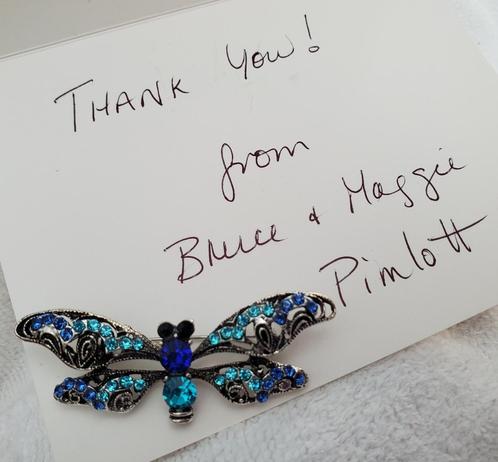 A handwritten thank you note rests on a soft, textured white surface, likely a blanket or towel. The note reads "Thank you!" at the top, followed by "from Bruce & Maggie Pimlott". A decorative dragonfly brooch sits at the bottom of the note. The brooch has a silver filigree design and is adorned with varying shades of blue gemstones.