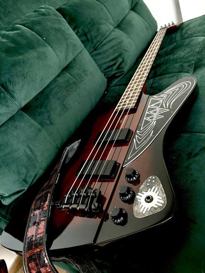 An electric bass guitar rests on a green velvet couch. The guitar features a unique design on its body and has several control knobs. The cozy couch background contrasts with the instrument's glossy finish.