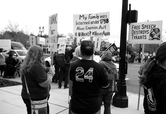 Black and white photo of a person holding a sign in the middle of a protest that says "Honk For Democracy" and another person holding a sign that says "My family was interned under the 1798 Alien Enemies Act - R U Next?  #NeverAgain". Another sign says "Free Speech Destroys Tyrants"