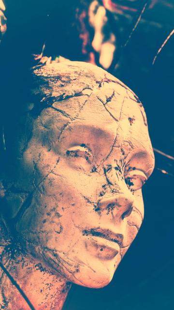 A cracked, weathered mannequin head with a solemn expression is captured in a dramatic play of light and shadow. The image evokes a sense of mystery and ancient artistry.