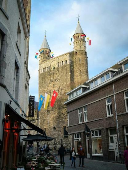 A picturesque street ,in Maastricht, scene featuring historic towers with flags, lined by modern buildings and an outdoor café. People can be seen walking and enjoying the ambiance.