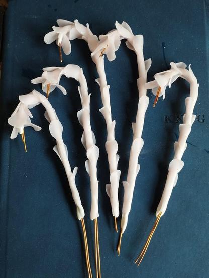 Five handmade Monotropa flower sculptures in progress, arranged on a dark blue surface. The pale, translucent stems and petals are sculpted with a delicate, waxy appearance. Each flower has visible copper wires extending from the base and some protruding from the flower tips, where the buds will be soldered later.