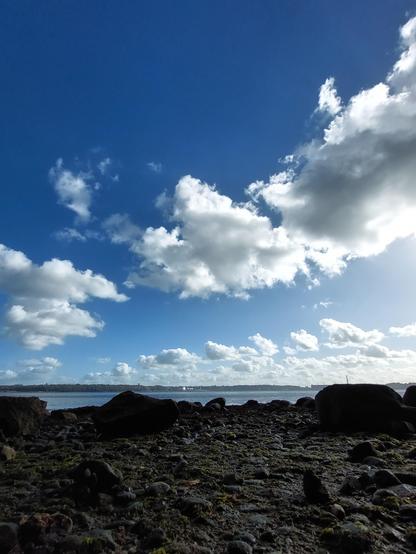 🇩🇪 Im Vordergrund sehr dunkle, fast schwarze Steine. An den Steinstrand anschließend blaues Meer. Im Hintergrund ein schmaler Streifen Land. Hoher blauer Himmel mit weißen Quellwolken. 
 🇬🇧 Dark, almost black stones in the foreground. Blue sea next to the stone beach. A narrow strip of land in the background. High blue sky with white spring clouds.