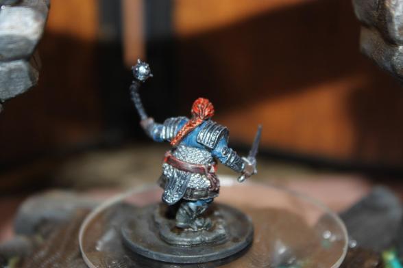 Alt text:
A rear view of a painted miniature depicting a red-haired warrior with a long braid, clad in blue and silver armor. The figure holds a spiked mace in one hand and a dagger in the other, standing on a textured base atop a clear display platform, framed by stone arch structures in the background.