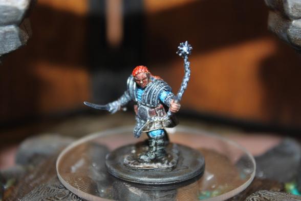 Alt text:
A painted miniature of a red-haired warrior stands on a round base, clad in blue and silver armor with a flanged mace raised in one hand and a curved dagger in the other. The figure is posed in a dynamic stance on a transparent platform, flanked by stone arch structures in the background.