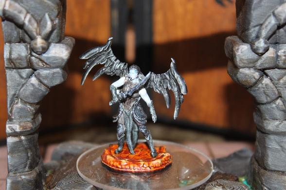 Alt text:
A painted miniature of a dark angelic figure with large, tattered black wings stands on a fiery orange base. The figure has pale blue-gray skin, long hair, and wears dark armor while gripping a sword diagonally across the chest. It is displayed on a clear platform between two stone pillars.