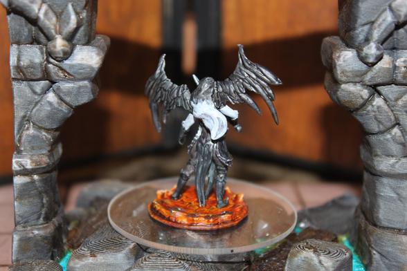 Alt text:
A rear view of a painted miniature depicting a dark-winged, angelic figure with outstretched black wings and flowing white hair. The figure stands on a fiery orange base, posed on a clear platform between two stone columns, with detailed armor and sculpted textures visible from the back.