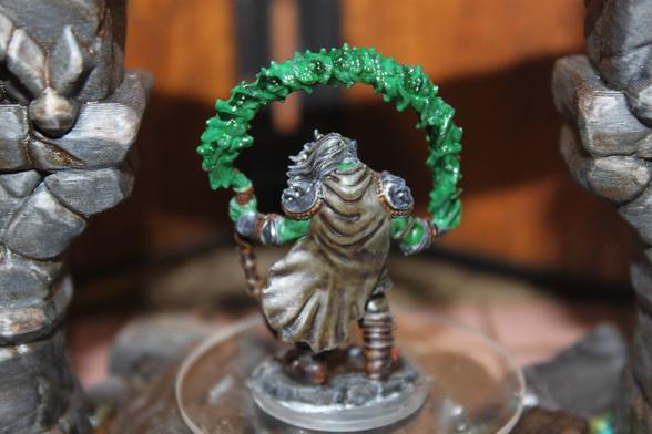 Alt text:
A rear view of a painted miniature depicting a green-skinned orc or half-orc druid holding a curved staff topped with a swirling green magical effect. The figure wears a weathered cloak and leather armor, with long hair flowing behind, standing on a rocky base between two stone columns.