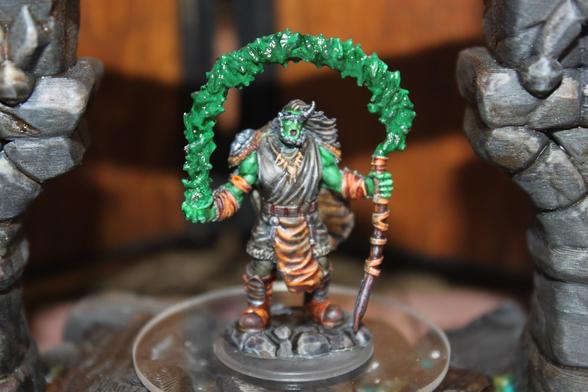Alt text:
A painted miniature of a green-skinned orc or half-orc druid stands on a rocky base, wielding a wooden staff that arches overhead with a swirling, translucent green magical effect. The figure is dressed in tattered robes and armor, with flowing hair and an intense expression, posed between two stone pillars.