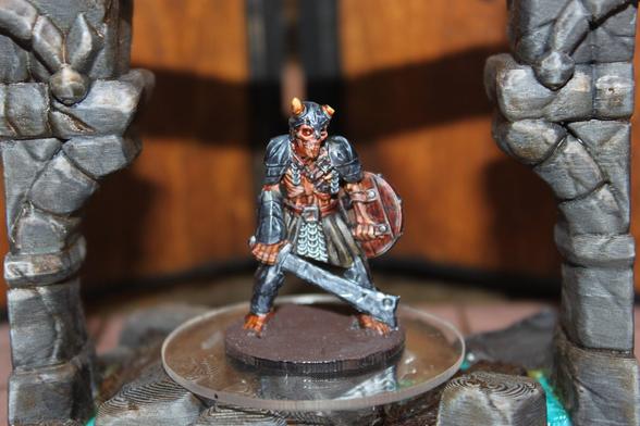 Alt text:
A painted miniature of a horned, skeletal warrior stands on a brown base. The figure is clad in dark armor with a scale-mail skirt, wielding a wide-bladed sword in one hand and a round shield in the other. It is posed between two stone pillars, with an expression of grim determination and weathered gear suggesting a veteran fighter or undead champion.