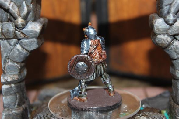 Alt text:
A rear view of a painted miniature depicting a horned, armored warrior with a muscular back and dark armor. The figure carries a round wooden shield and a large sword, and stands on a brown base placed atop a clear platform between two stone columns.