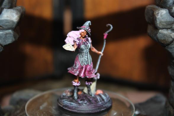 Alt text:
A painted miniature of a female spellcaster in a whimsical pink and purple outfit. She wears a pointed hat adorned with flowers, has long curly pink hair, and holds a curved staff in one hand while presenting a tray of pastries in the other. The figure stands on a textured base between two stone pillars, evoking a charming and fantastical vibe.