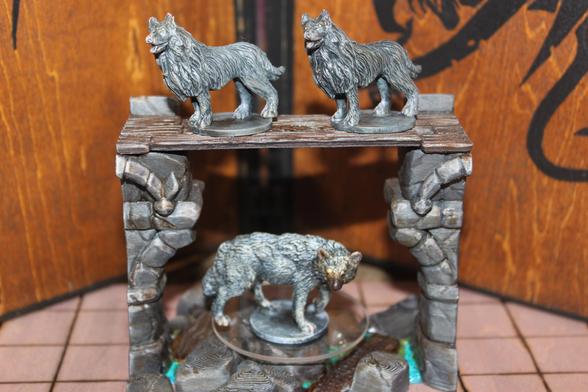 Alt text:
A group of three painted wolf miniatures posed in a dungeon-like diorama. Two majestic, long-maned wolves with raised heads stand side-by-side on a wooden platform supported by stone pillars. Below them, on the ground level, a more realistically textured gray wolf with glowing eyes prowls cautiously atop a circular base. The backdrop features a wooden wall with black draconic designs.