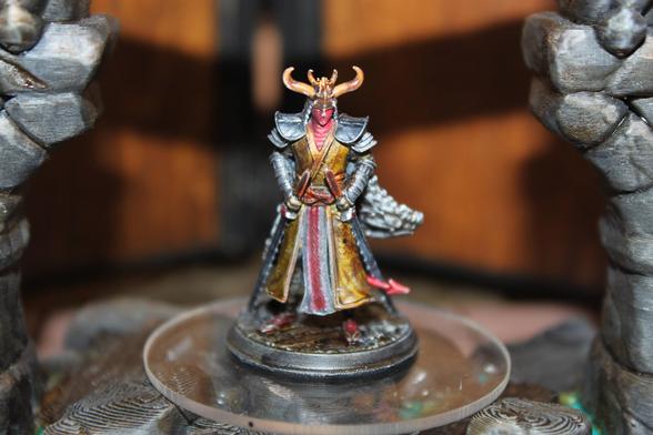 Alt text :
A painted miniature of a tiefling samurai, adorned in ornate crimson and gold robes with dark armored pauldrons. The figure wears a horned helm and has a flowing textured cloak draped behind. Twin swords are sheathed at the waist, and the tiefling’s stance is calm yet commanding, posed atop a circular base within a stone-pillared display setting.