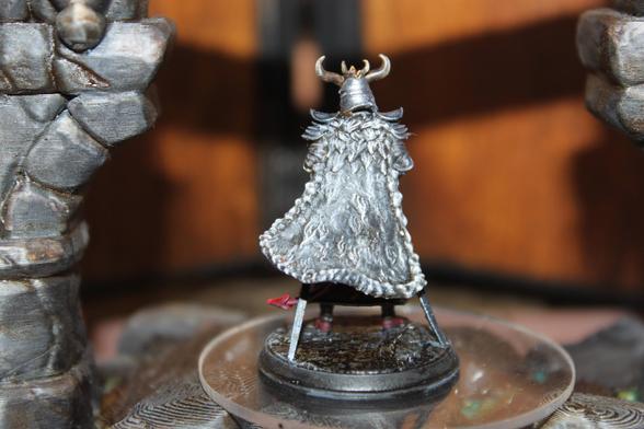 Alt text:
Rear view of a painted tiefling samurai miniature wearing a large, flowing gray fur cloak with intricate texturing. The figure's horned helmet is visible above the cloak, and twin red-tipped tails peek out from beneath the garment. The miniature stands poised on a round base within a stone-arched display setup.