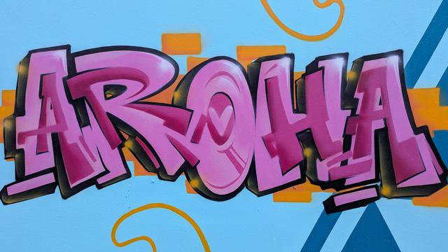 "Aroha" written as street art in large patterned pink letters on a sky blue background