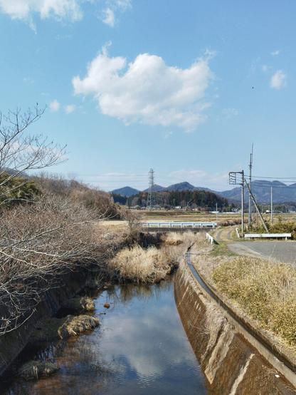 A creek straightened by concrete embankments curves slightly to the right. There are barren shrubs to the left and fields on the right. In the background are small mountains.