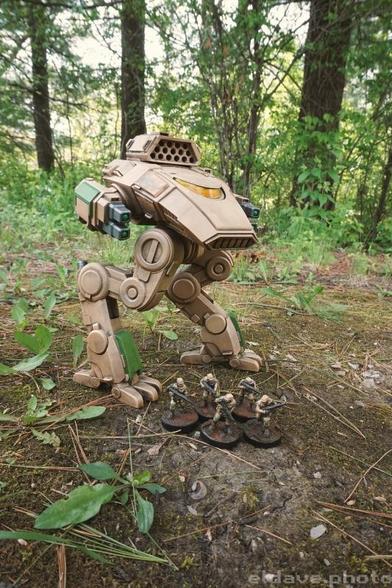 A 3d printed Battletech model from Locust Labs, scaled up 600%, and standing outside in the woods with 5 human soldiers standing around its feet.