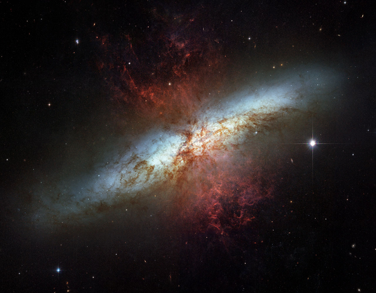 Image of Messier 82 -- a starburst galaxy. The galaxy has a bright central region and glowing red and blue clouds of gas extending outward, surrounded by scattered stars and a dark background.