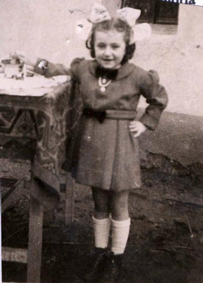 A young girl in a dress standing next to a high table. She has two large bows on in her hair.
